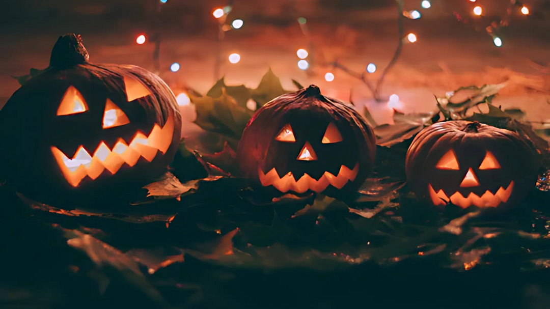 Three jack-o-lanterns glowing in the dark, surrounded by autumn leaves and festive string lights in the background.