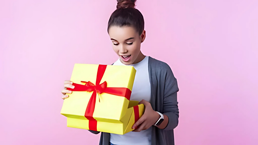 A tween girl with a top bun, wearing a gray cardigan and white shirt, looks excitedly at a yellow and red gift box she is opening. The background is pink.