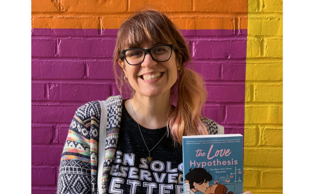 Author Ali Hazelwood smiling and holding a copy of her book 'The Love Hypothesis' in front of a colorful brick wall