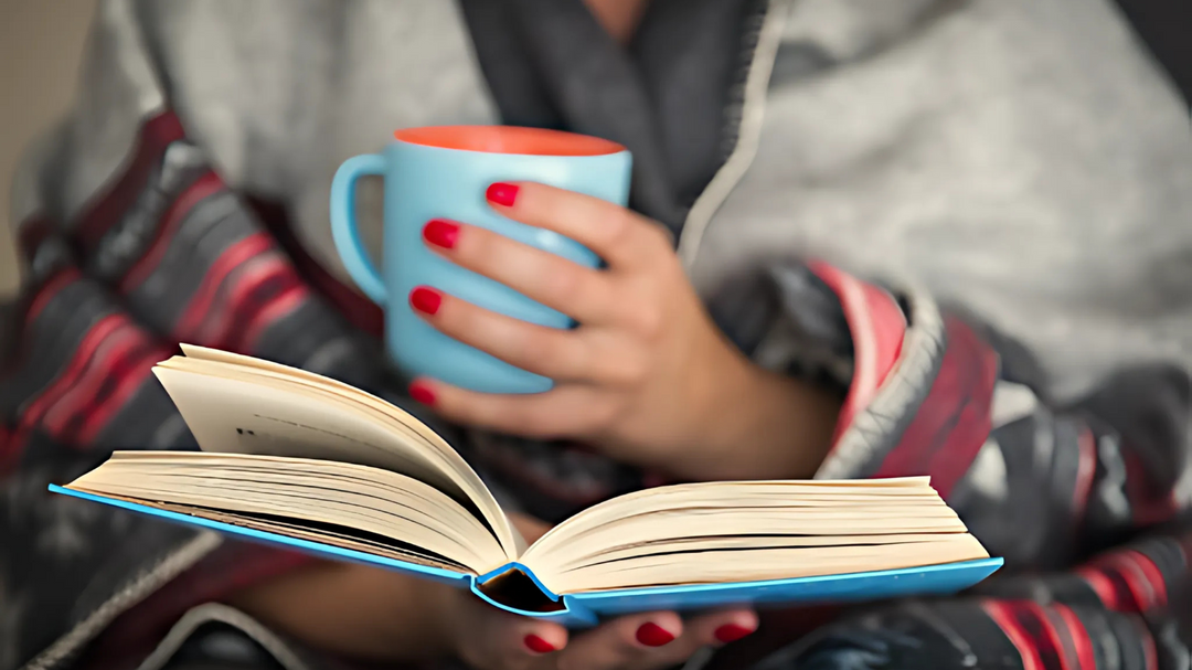 A person wrapped in a warm blanket holds a blue mug while reading a book, creating a relaxing and cozy atmosphere.