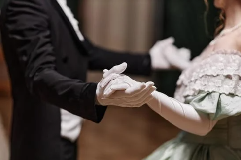 A close-up of two individuals dancing at a formal event, dressed in elegant attire, holding hands while wearing white gloves.