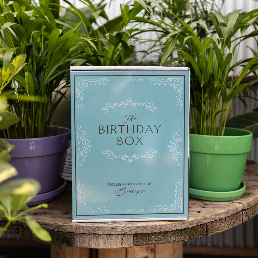 A blue Birthday Box sits on a table outside surrounded by various color pots and potted plants