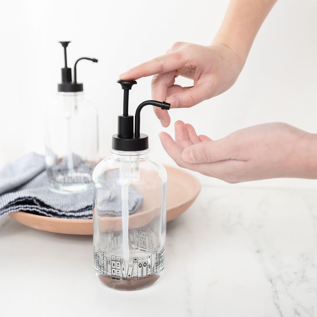 A set of two glass soap dispensers sit on a white marble countertop. A pair of white hands presses on the pump to dispense liquid. The glass bottles are rimmed with a book stack along the bottom of each jar.