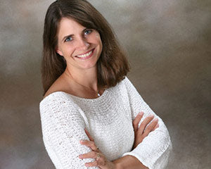 Author Alix Rickloff is a middle-aged white woman with shoulder-length brown hair. She stands with her arms crossed against a grey-brown background looking up toward the camera. She wears a small pendant necklace and a white sweater.