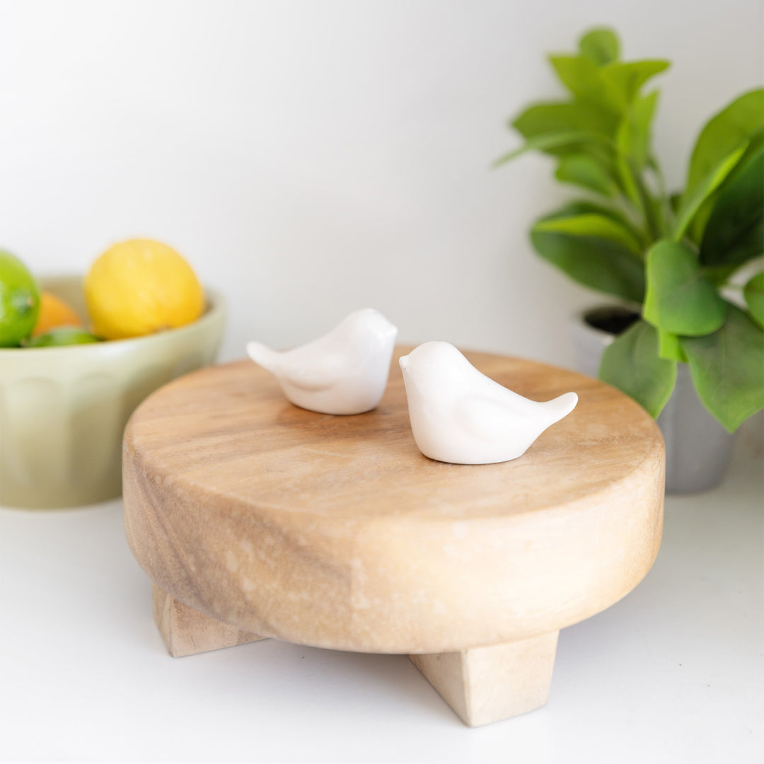 a set of two small white salt and pepper shakers that are shaped like birds sit on a piece of wood. There is a plant and a bowl of fruit in the background