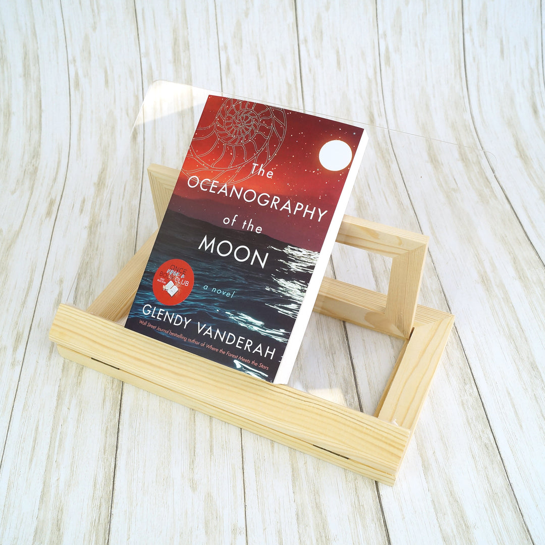 a light wood and plexiglass book stand holds a paperback book.