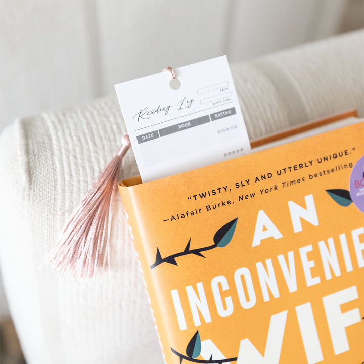 A reading log bookmark sticks out of the top of an orange hardcover book. The columns (including Date, Book, and Rating) are clearly visible. The tassel is tucked behind the bookmark.