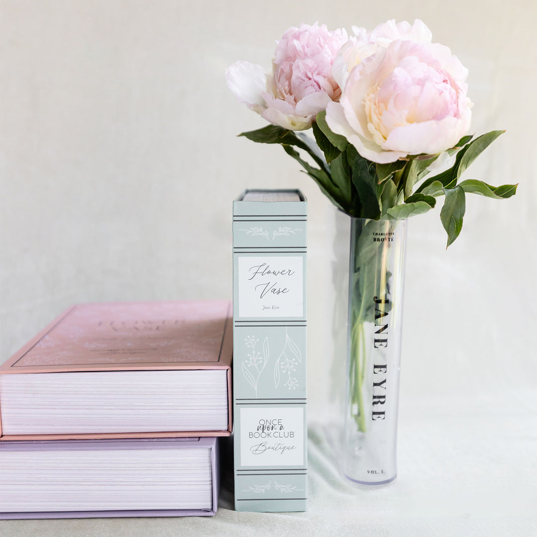 Two Once Upon a Book Club Boutique boxes lay on their side with the printed edges of the boxes facing the camera. One box is standing up to show how much it looks like a book. The spine of the box reads "Flower Vase, Jane Eyre". Standing next to the box is the book-shaped vase designed to look like a copy of Jane Eyre. It holds light pink flowers and greenery.