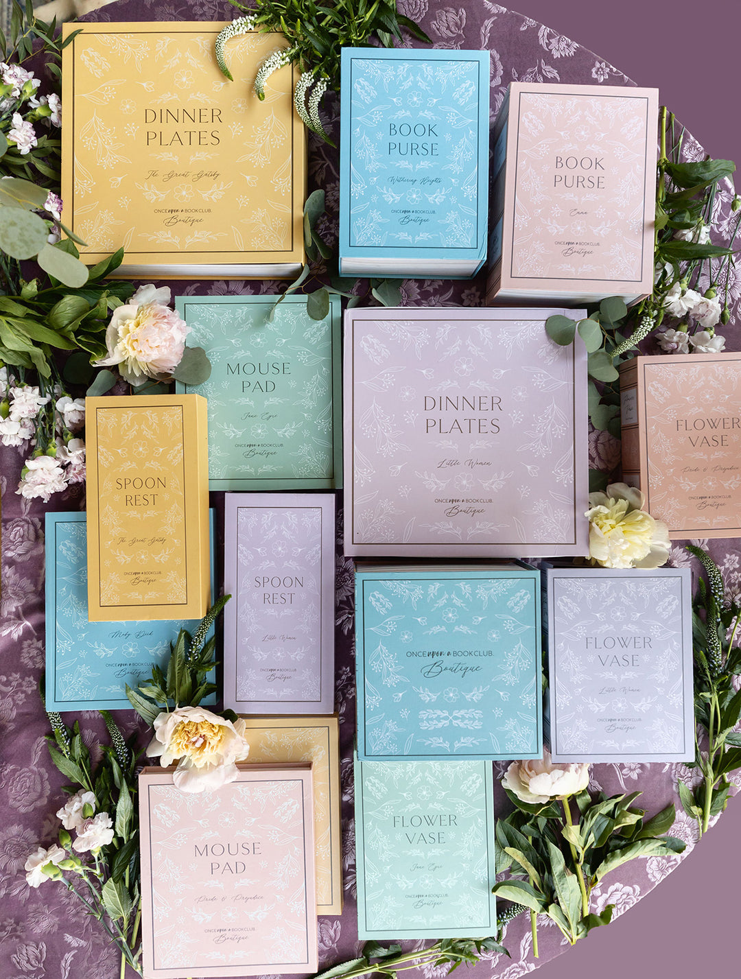 A collection of various colored Once Upon a Book Club Boutique boxes sits on a purple floral background. The boxes are each printed to contain various products including purses, plates, mouse pads, spoon rests, and vases. The boxes are surrounded by greenery.