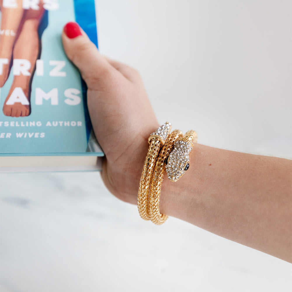 a woman's hand holds a book. on the wrist is a gold cobra bracelet with a crystal head and black eyes