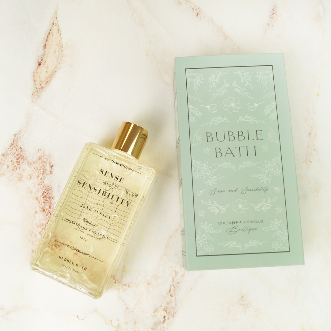 A bottle of bubble bath with a gold cap lays on a pink marbled surface. Next to the bubble bath is a green box labeled "Bubble Bath" "Sense and Sensibility" "Once Upon a Book Club Boutique"