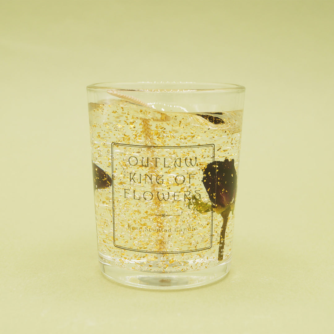 A glass candle votive containing jelly wax, gold glitter, and rose petals. The jar reads "Outlaw King of Flowers" Rose Scented Candle.