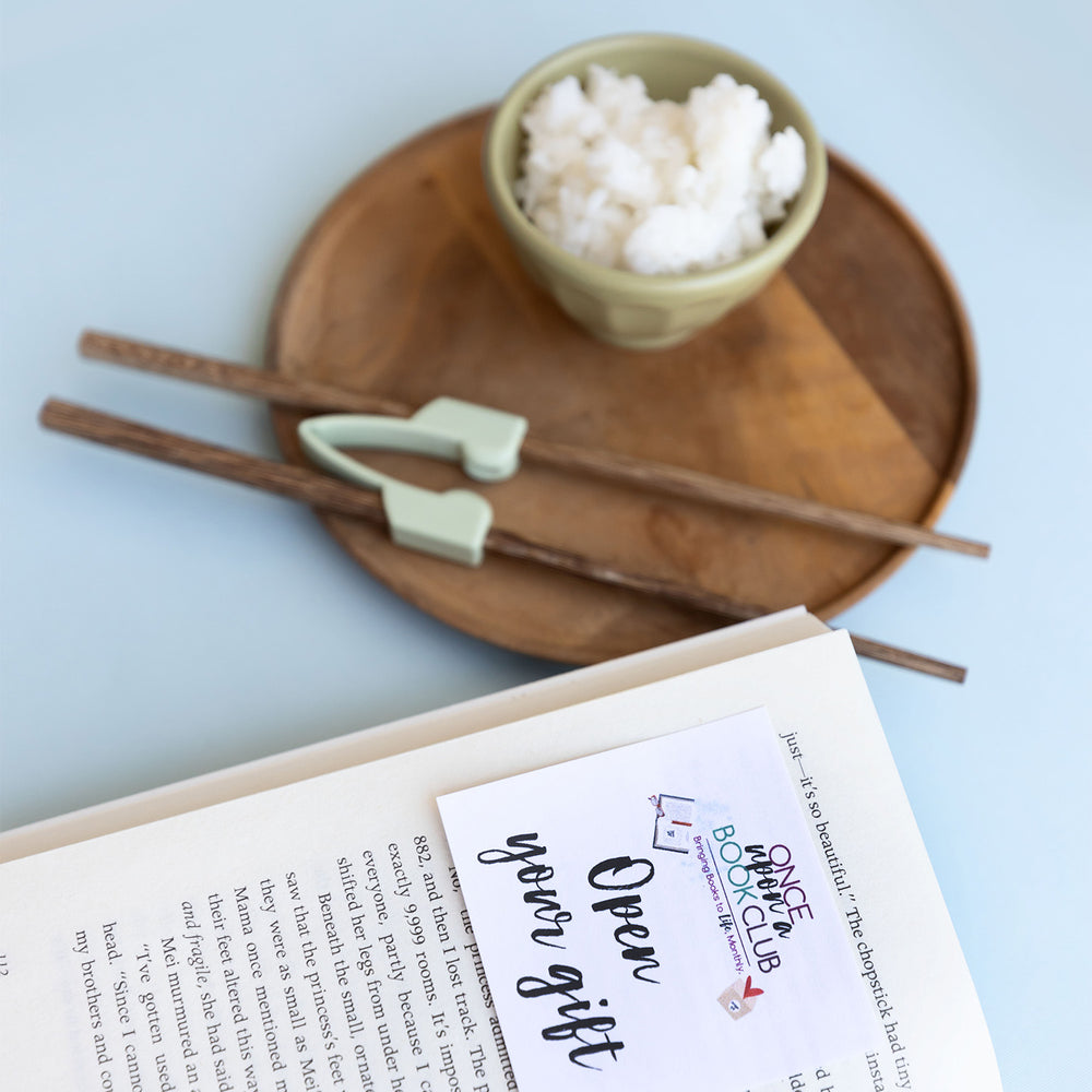 a pair of training chopsticks sits on a wooden plate next to a bowl of rice and an open-your-gift sticky note stuck inside the book.