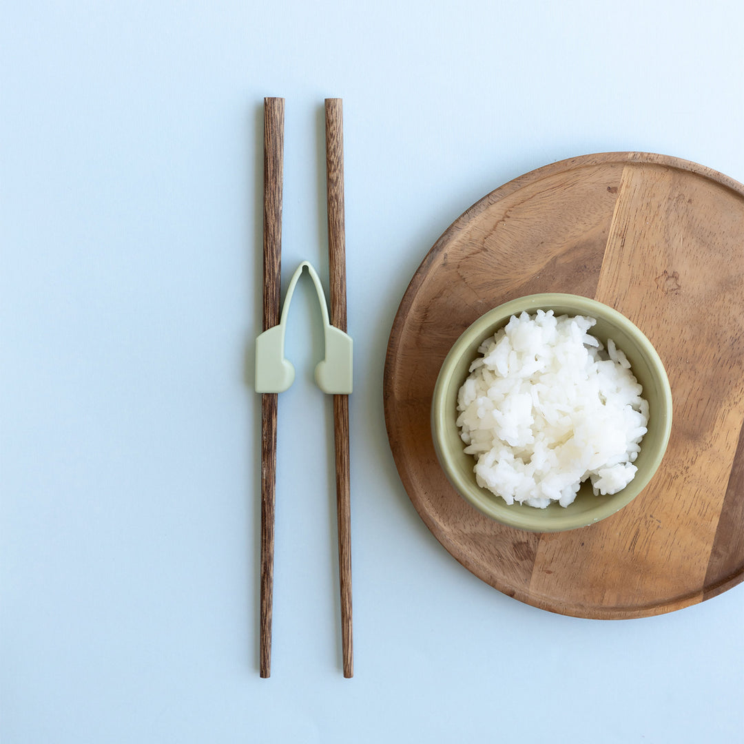 a pair of training chopsticks sits next to a bowl of rice on a wooden tray.