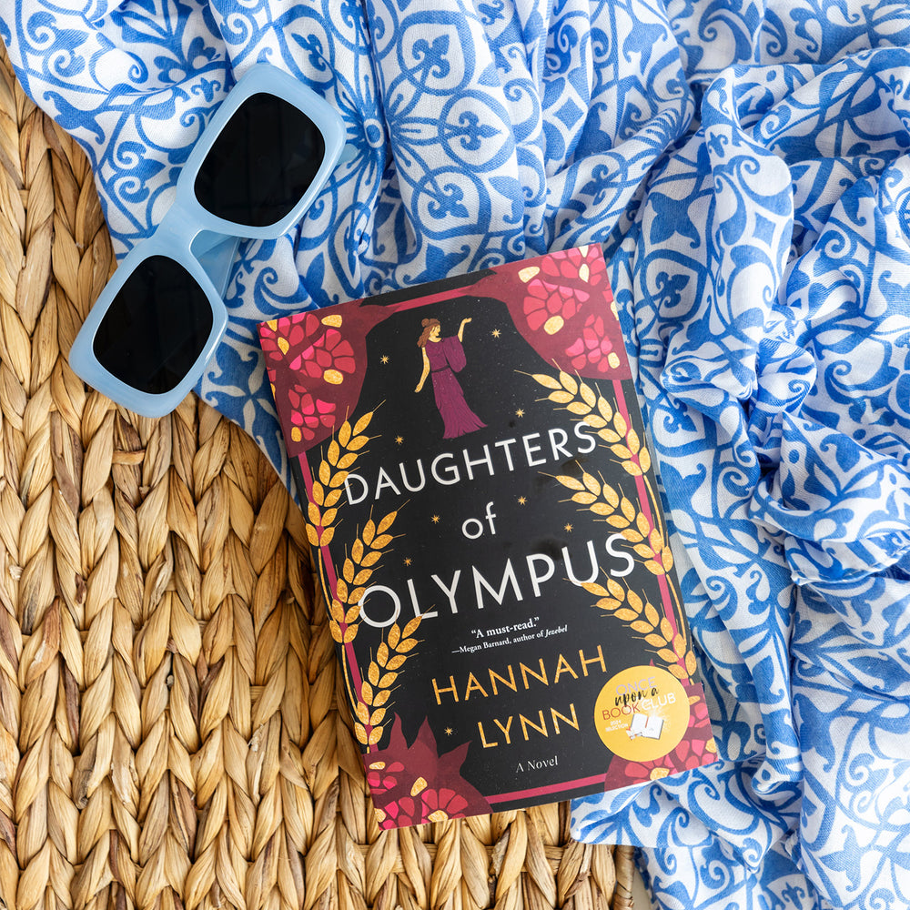 a paperback copy of Daughters of Olympus by Hannah Lynn sits on a wicker background surrounded by a scarf and sunglasses.