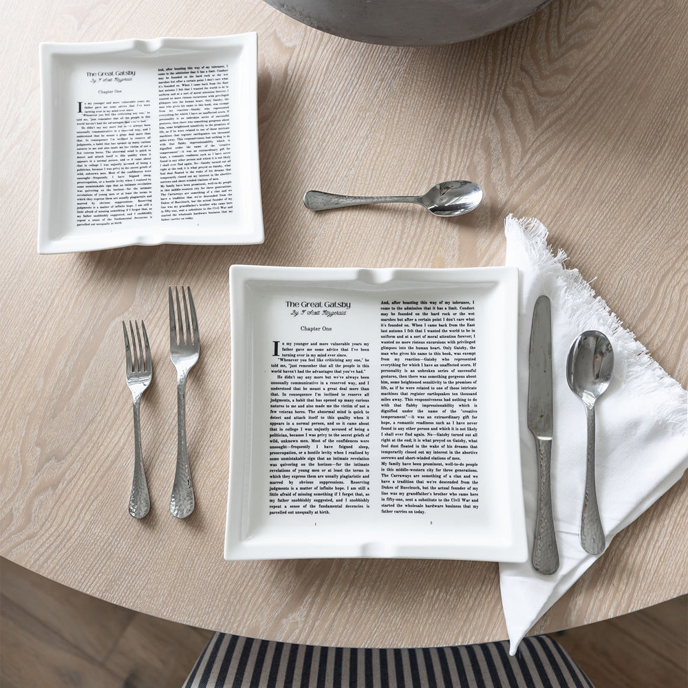 A flat lay image featuring dinner plates shaped like books inspired by The Great Gatsby by F. Scott Fitzgerald. The large and small plate are both visible on a wooden table. Silverware is placed around the plates in a standard table setting format. A white cloth napkin sits to the right of the plate under the knife and soup spoon.