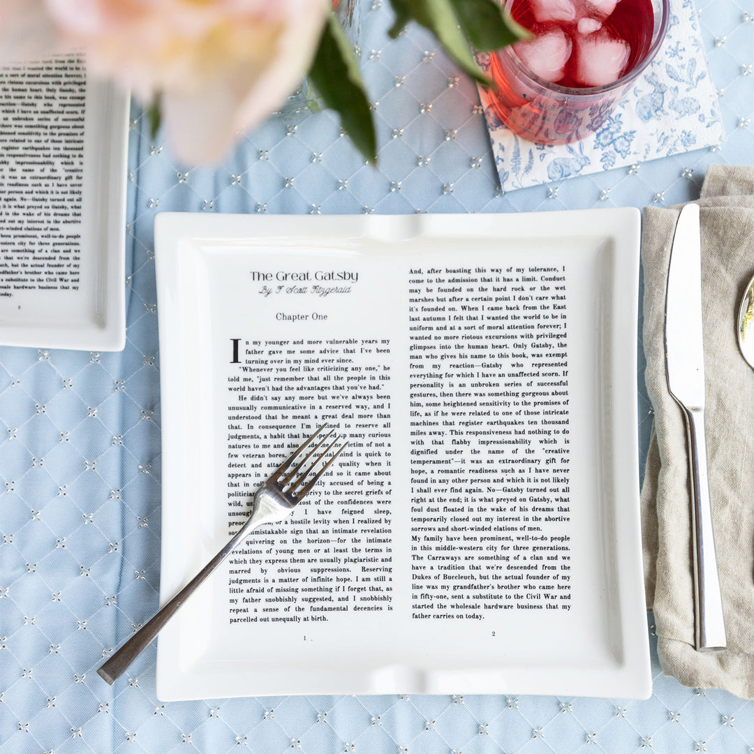A flatlay image featuring dinner plates shaped like books inspired by The Great Gatsby by F. Scott Fitzgerald. The plates sit on a blue patterned tablecloth. A floral arrangement is blurred at the top of the image. A glass filled with a red beverage sits to the top right of the image on a blue and white floral napkin. A silver fork sits across the large plate. A silver knife and spoon sit to the right of the large plate on a grey cloth napkin.