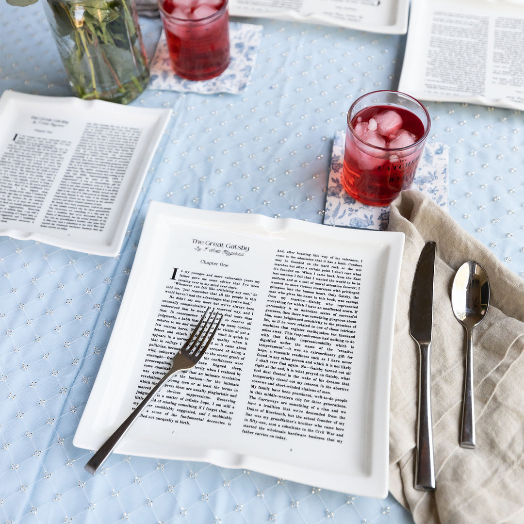 A flat lay image featuring dinner plates shaped like books inspired by The Great Gatsby by F. Scott Fitzgerald. The plates sit on a blue patterned tablecloth. Both the large and small plates are visible. Silverware is placed around the plates. A drinking glass filled with a red beverage can be seen at two table settings from this angle. A grey napkin sits to the right of the large plate holding the knife and spoon.