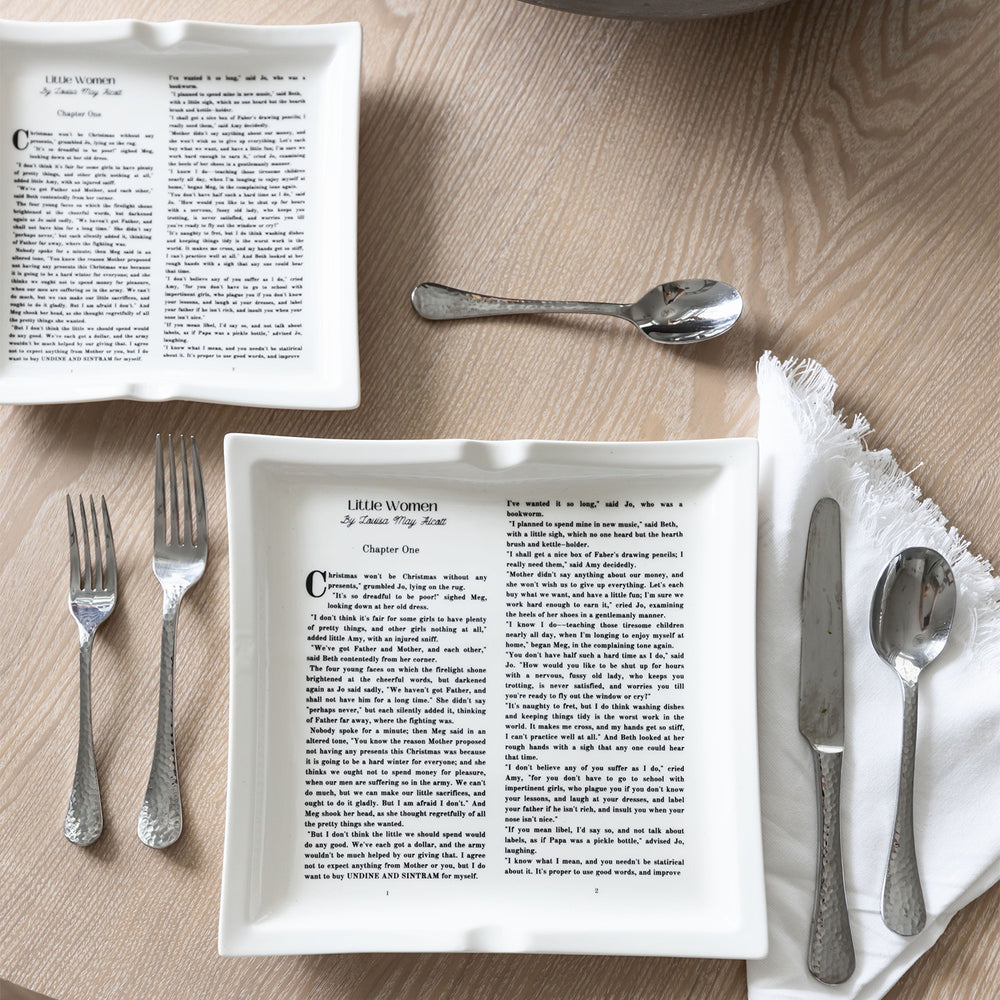 A flat lay image featuring dinner plates shaped like books inspired by Little Women by Louisa May Alcott. The large and small plates are visible on a wooden table. Silverware is spread next to the plate set, some of which sits to the right of the large plate on a white cloth napkin.