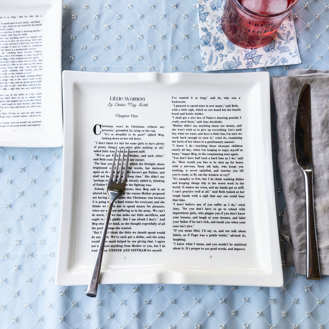 A flat lay image featuring dinner plates shaped like books inspired by Little Women by Louisa May Alcott. The plates sit on a light blue patterned tablecloth. A silver fork is placed across the plate. A silver knife sits to the right of the plate on a grey cloth napkin. A glass filled with a red colored beverage sits at the top of the image on a blue and white floral napkin.