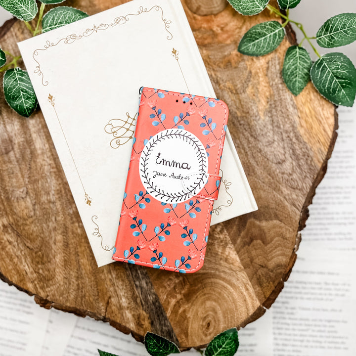The front of a phone case inspired by Emma by Jane Austen. The phone case sits on a white book cover on a wood slice surrounded by greenery.