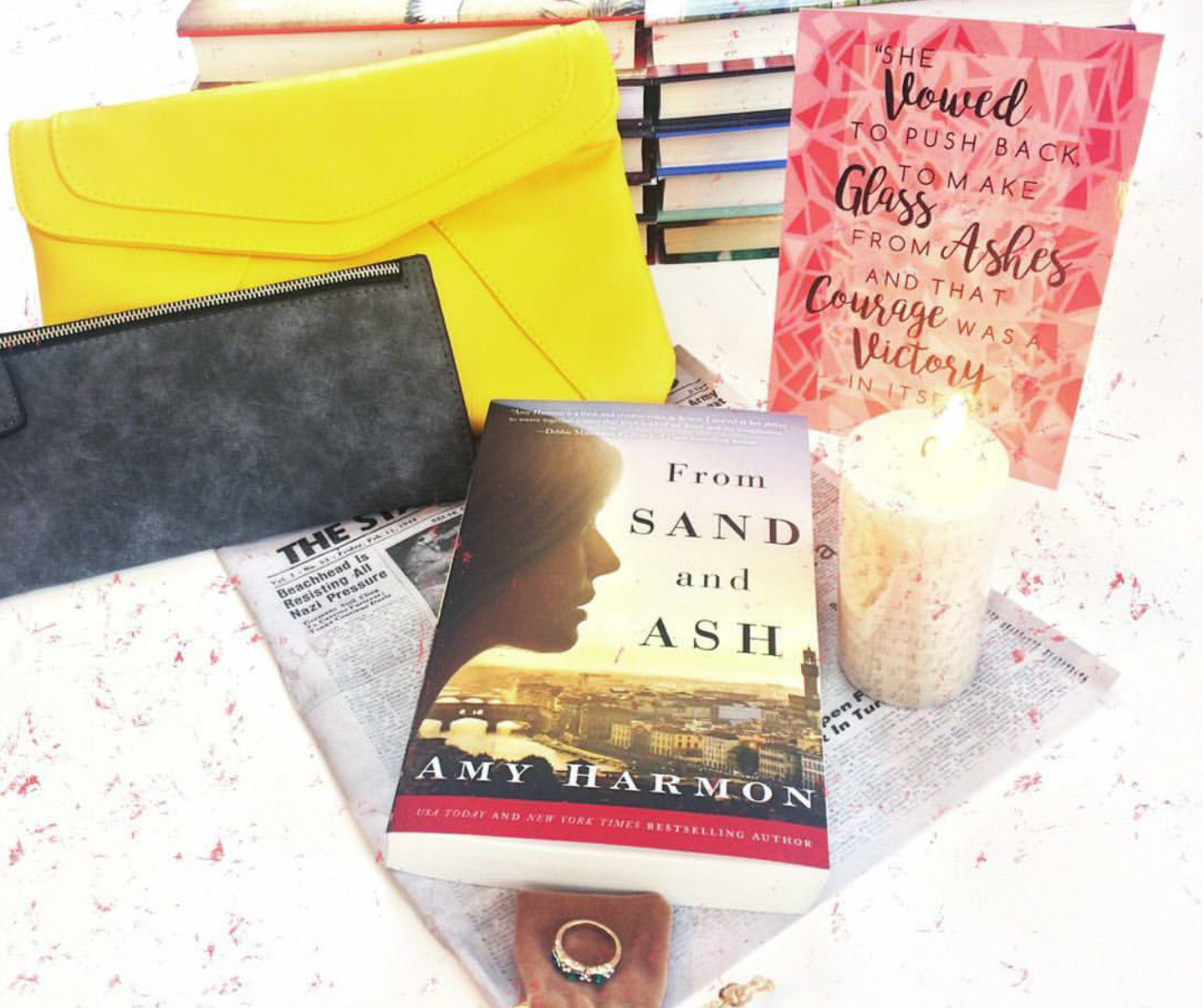 A paperback copy of From Sand and Ash by Amy Harmon sits on top of a set of unwrapped gifts inspired by the novel. These include a grey zippered wallet, a yellow crossbody purse, a candle printed with music notes, a ring, and a newspaper article. 