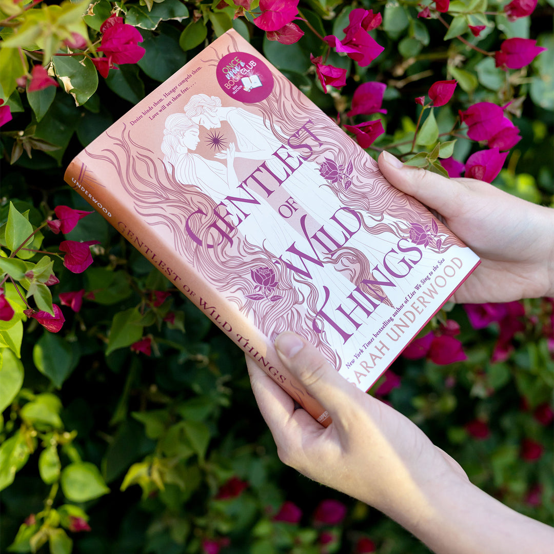 a hardcover copy of Gentlest of Wild Things by Sarah Underwood outdoors being held up in front of a flowering bush.