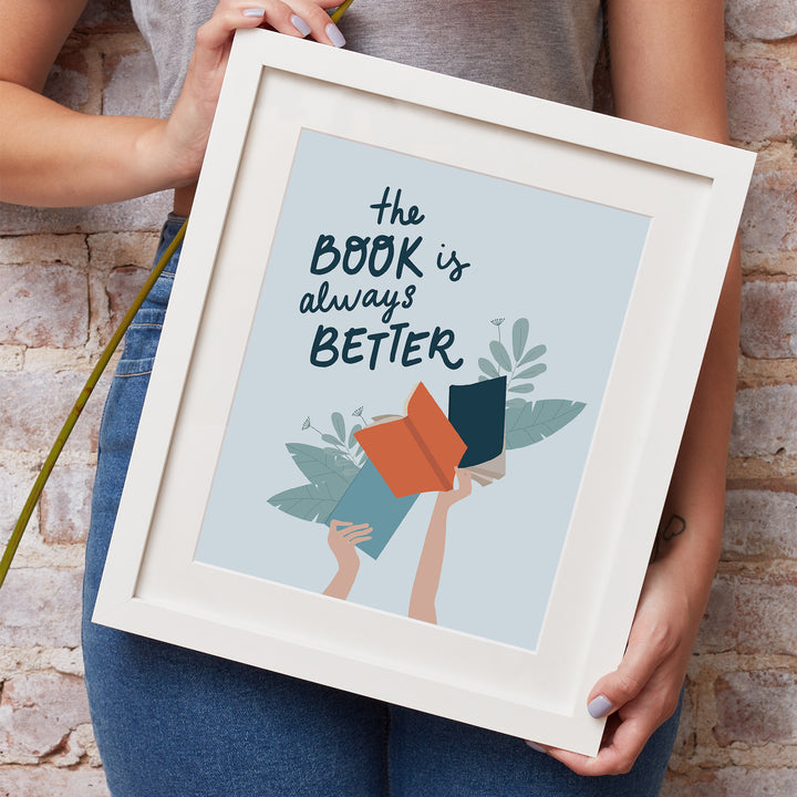 A white woman's hands hold a white matted frame showcasing the "The Book Is Better" art print. She is wearing jeans and a grey shirt and leaning against a brick wall.