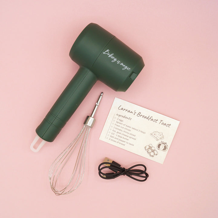 a wireless, dark green hand mixer with whisk attachment, charging cables, and a recipe card sits on a pink background. The mixer has the words "Baking is Magic" printed on the top barrel.