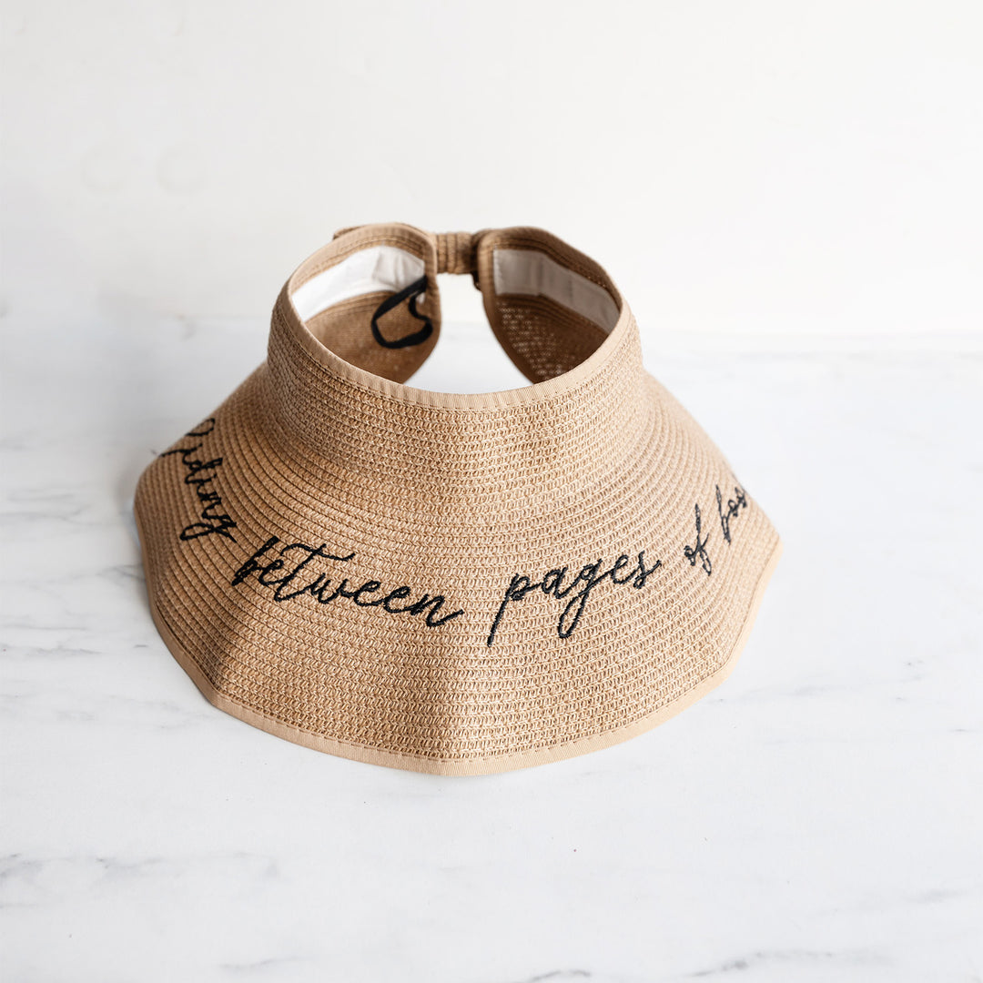 An open top, sand-toned sun hat featuring an embroidered quote on the brim of the hat: hiding between pages of books