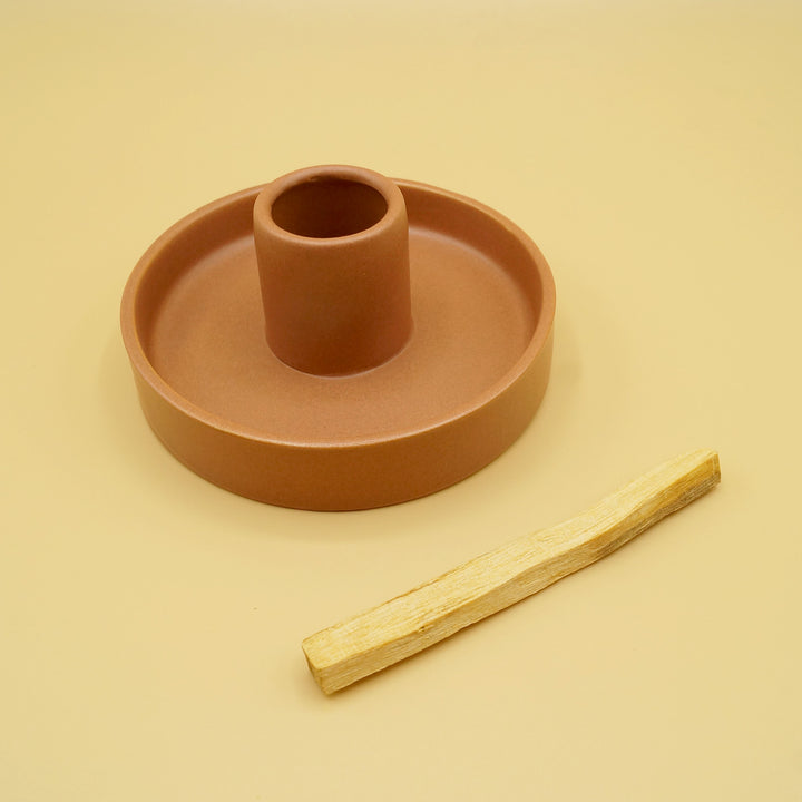 A clay colored circular dish with a large hole in the center stands about 2 inches tall. A wooden palo santo stick sits next to the holder. The stick would be inserted in the middle of the holder and lit on fire to release pleasant smelling smoke.