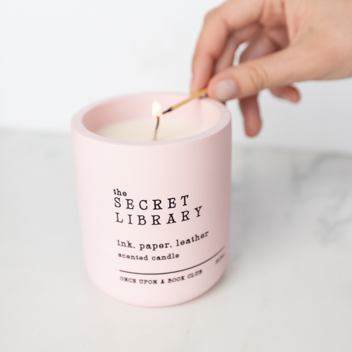 a white hand holds a match to a pink ceramic candle reading 'The Secret Library. ink. paper. leather. scented candle.' in black text.