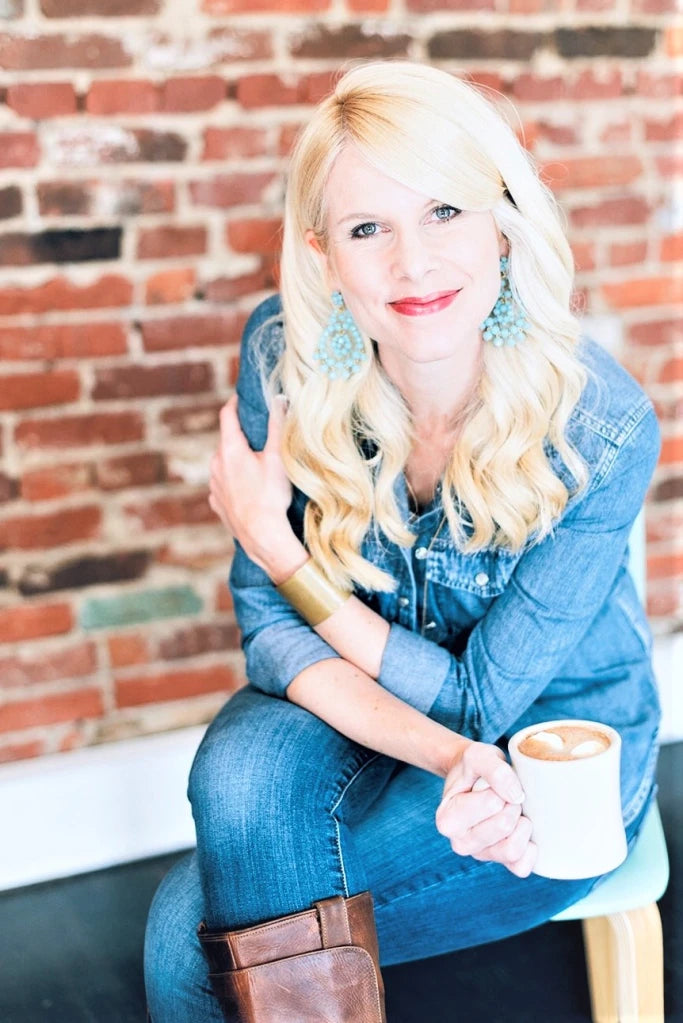 Author Kristy Cambron is a middle-aged white blonde woman wearing a jean jacket and jeans. She holds a cup of coffee in front of a brick wall.