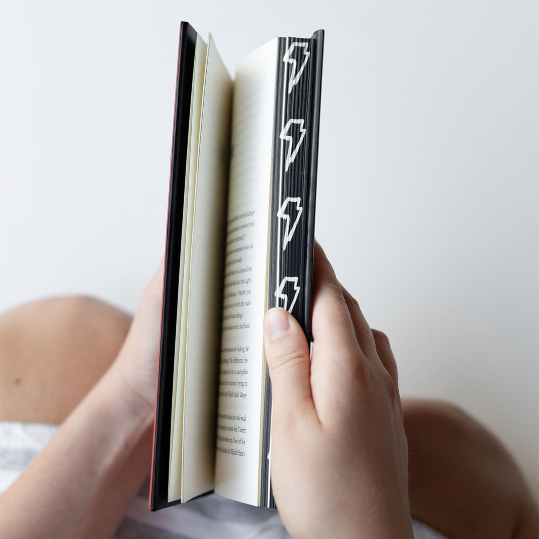 hands flip the pages of the custom edition of The Lightning Bottles by Marissa Stapley showing off the stenciled page edges featuring a black background and a white lightning bolt design.