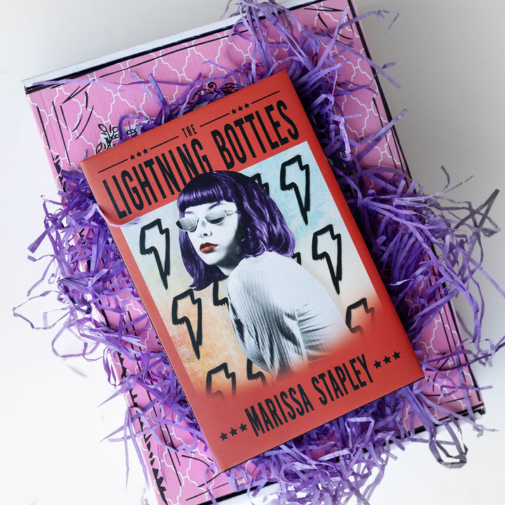 a custom hardcover edition of The Lightning Bottles by Marissa Stapley sits on purple easter grass on a pink box.