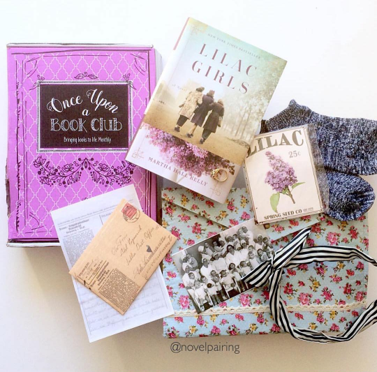 A hardcover copy of Lilac Girls by Martha Hall Kelly sits on a pink Once Upon a Book Club box. Surrounding it are the unwrapped gifts including a pair of socks, a packet of lilac seeds, a fabric envelope clutch, a headband, a photo, and a letter.