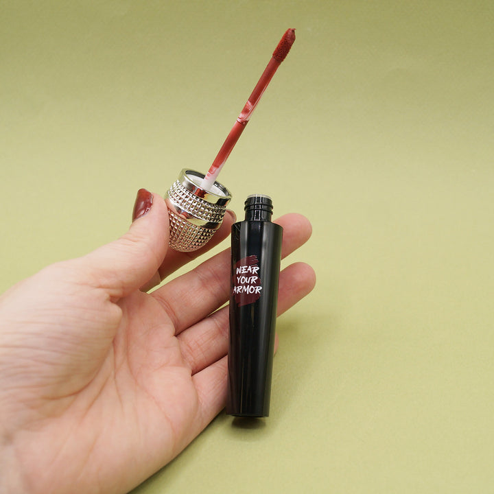 a white hand holds a black microphone-shaped tube that says Wear Your Armor. the top of the microphone tube is off and shows the color of the liquid lipstick inside is a maroon color.