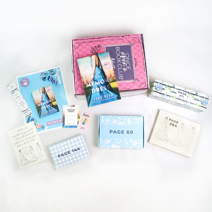 A paperback copy of The Memory Dress by Jade Beer sits on a pink Once Upon a Book Club box surrounded by wrapped gifts labeled with coordinating gift wrap and corresponding paper items.