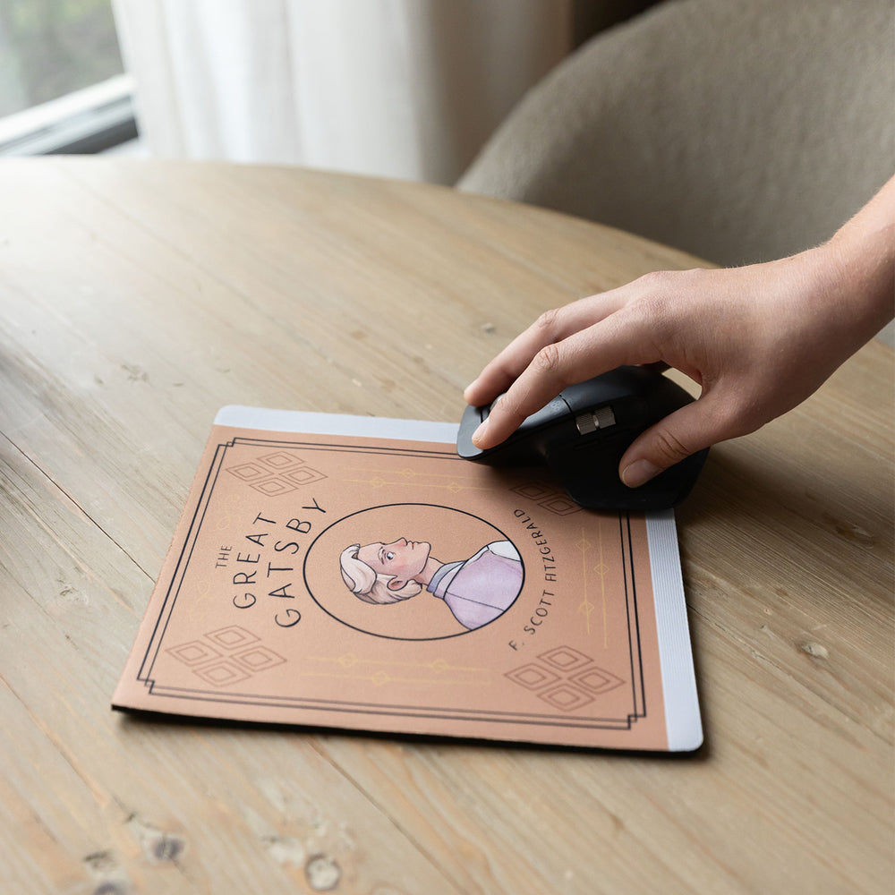 A light orange mouse pad meant to look like the book cover of The Great Gatsby featuring an illustration of Jay Gatsby in the center. The mousepad sits on a wooden table. A white hand grips a black computer mouse toward the bottom of the mouse pad.