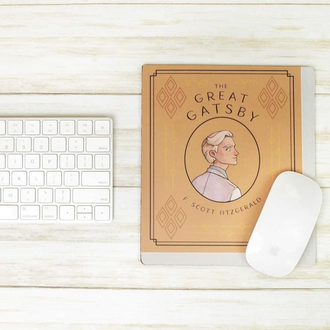 A light orange mouse pad meant to look like the book cover of The Great Gatsby featuring an illustration of Jay Gatsby in the center. On a white wooden background. A computer keyboard sits to the left of the mousepad and a computer mouse sits on the bottom right of mousepad.