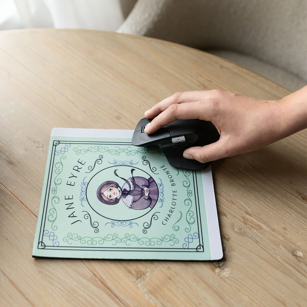 A light green mouse pad meant to look like the book cover of Jane Eyre featuring an illustration of Jane in the center. A white hand holds a black computer mouse on the mouse pad on a wooden table.