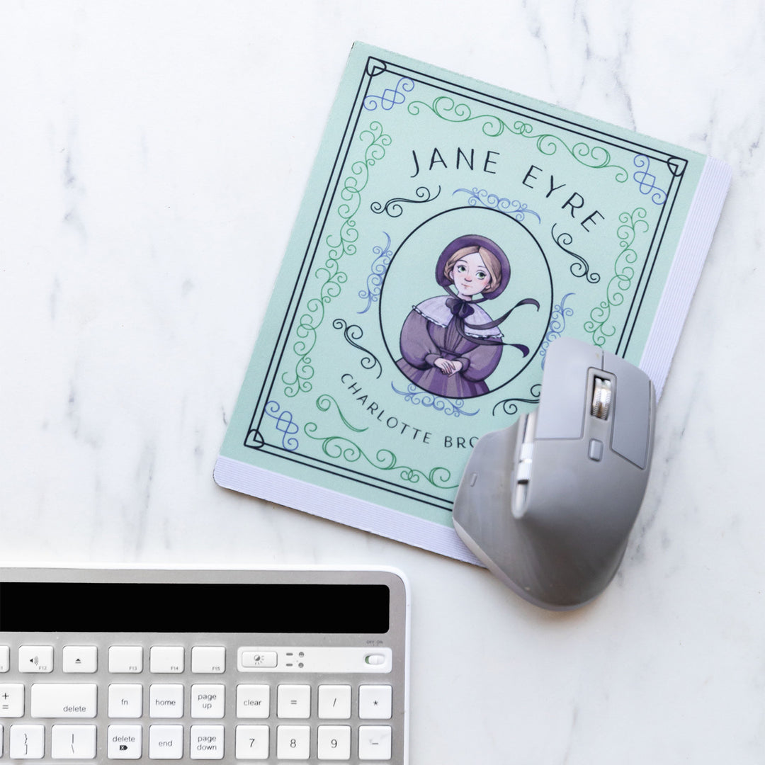 A light green mouse pad meant to look like the book cover of Jane Eyre featuring an illustration of Jane in the center. The mousepad sits on a white marble surface with a grey computer mouse on the lower right corner and keyboard visible toward the bottom of the image.
