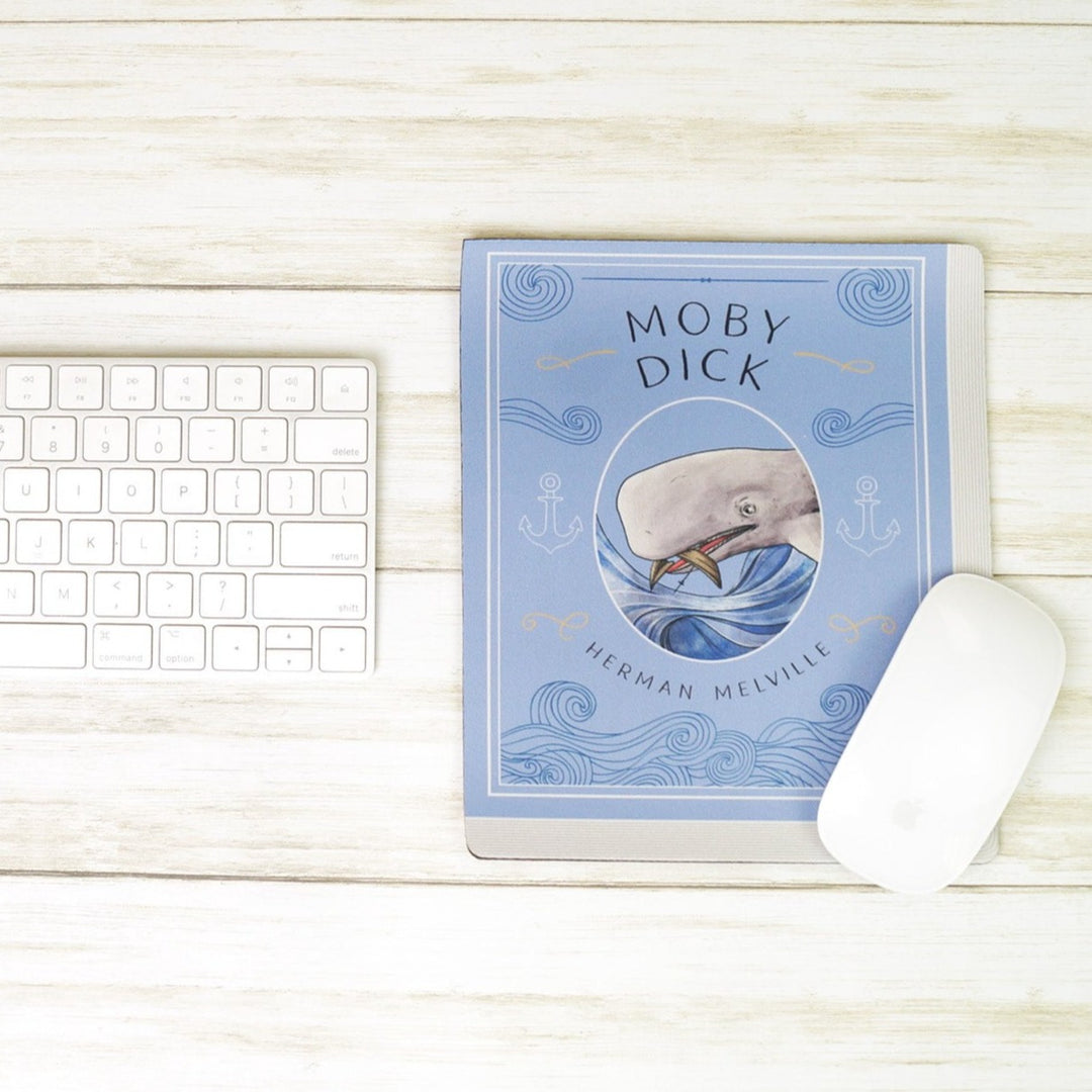 A light blue mouse pad meant to look like the book cover of Moby Dick featuring an illustration of the whale, Moby Dick, in the center. The mousepad sits on a white wooden table. A keyboard sits to the left of the mousepad and computer mouse sits on the lower right corner of the mouse pad.
