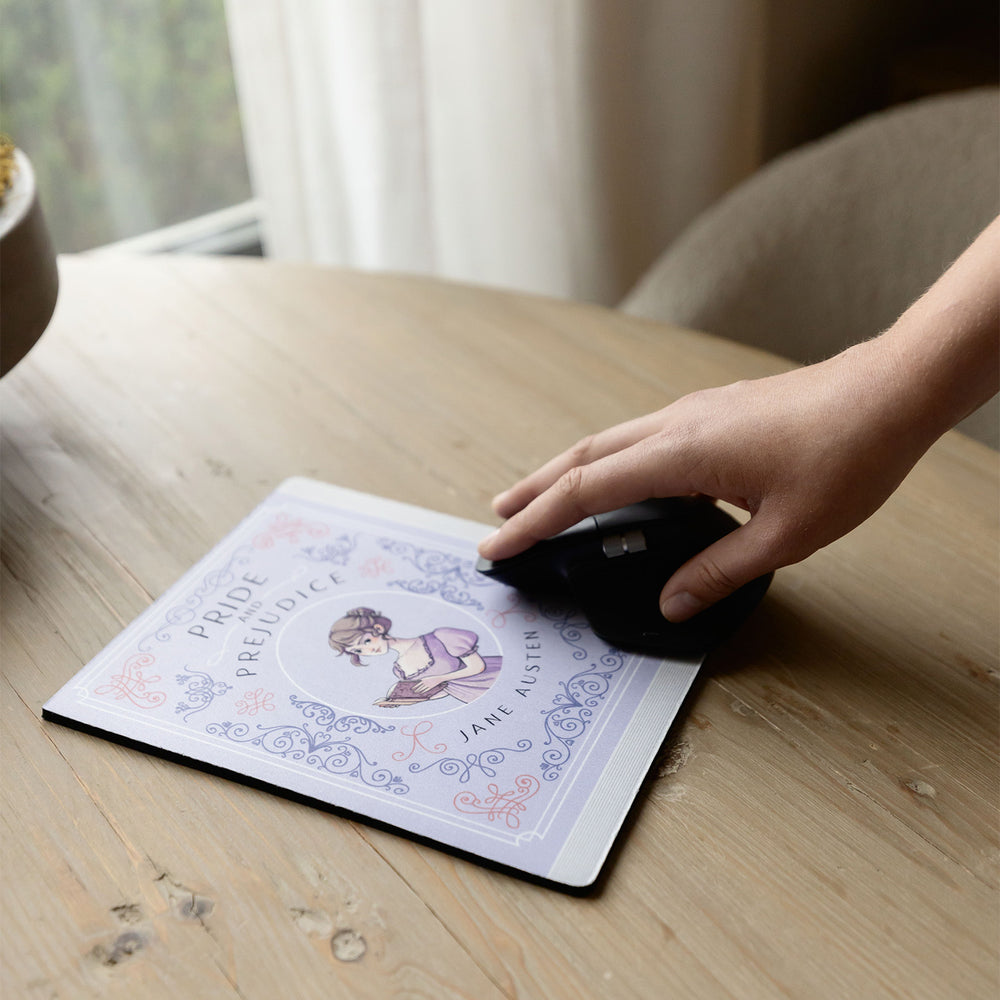 A light purple mouse pad meant to look like the book cover of Pride and Prejudice by Jane Austen featuring an illustration of Elizabeth Bennet in the center. The mousepad sits on a wooden table. A white hand grips a black computer mouse toward the bottom of the mouse pad.