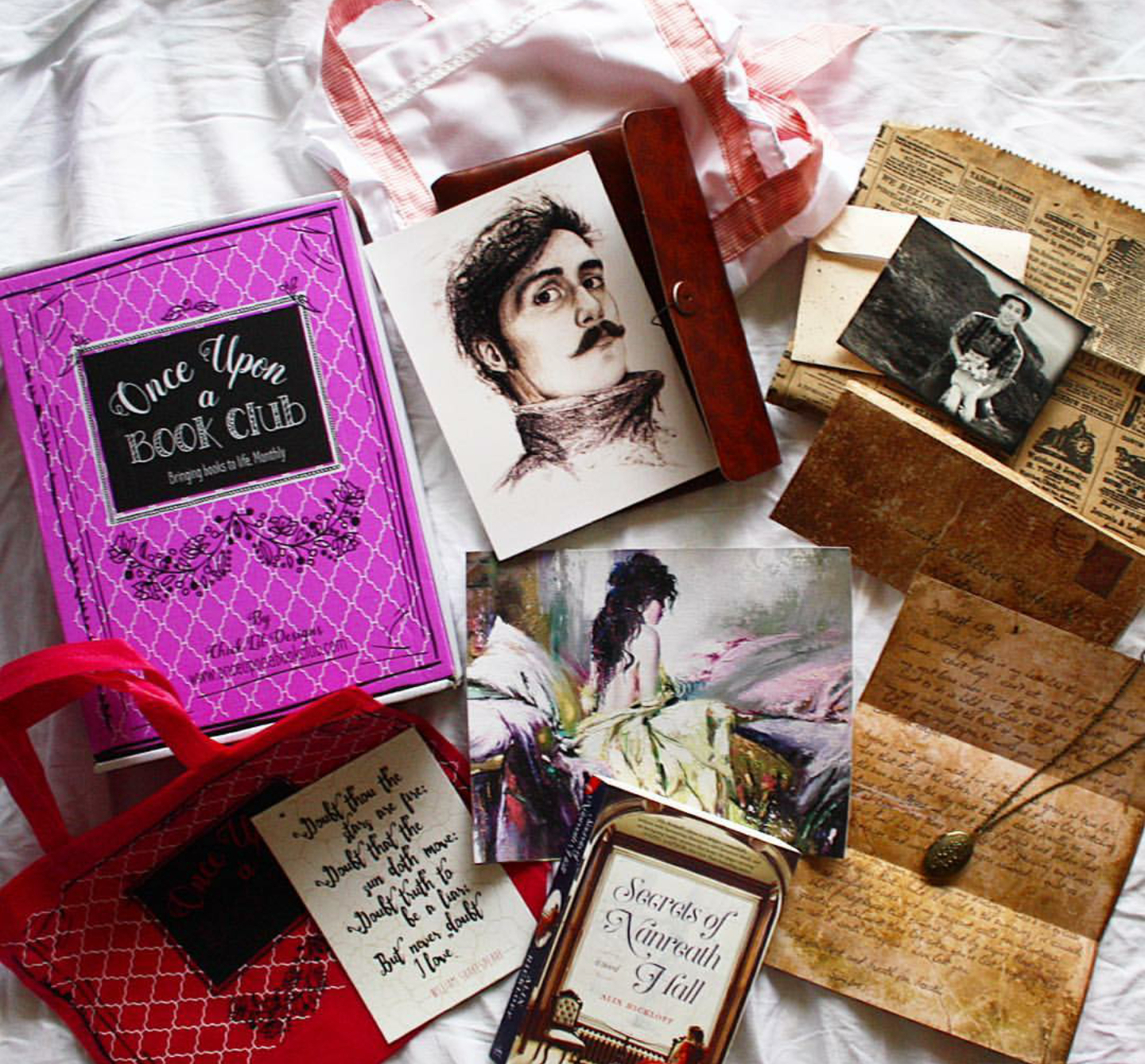 A paperback copy of Secrets of Nanreath Hall by Alix Rickloff sits next to a pink Once Upon a Book Club box. Surrounding the book and box are unwrapped gifts pulled from the story, including a faux leather portfolio with a sketch of a mustached man, a letter, a locket, an envelope containing a photograph, an apron, and a watercolor painting.