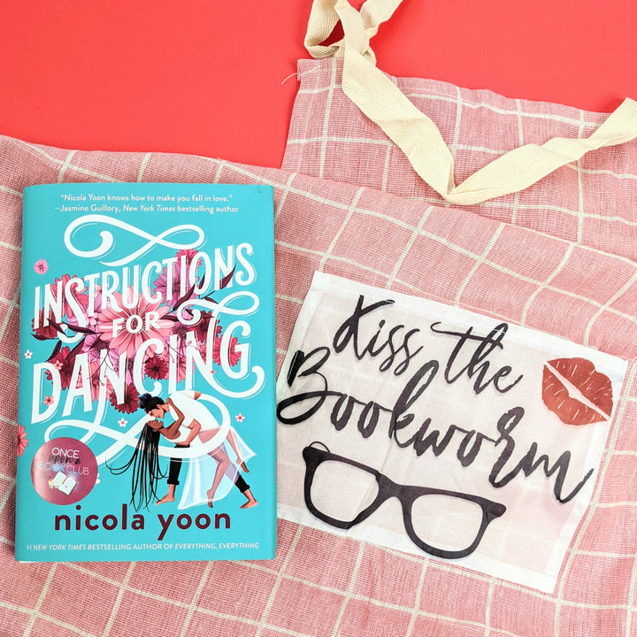 the pink gingham apron featuring a pocket with the words "Kiss the Bookworm" and a pair of glasses and lipstick kiss mark. With a copy of Instructions for Dancing by Nicola Yoon. On a red background.