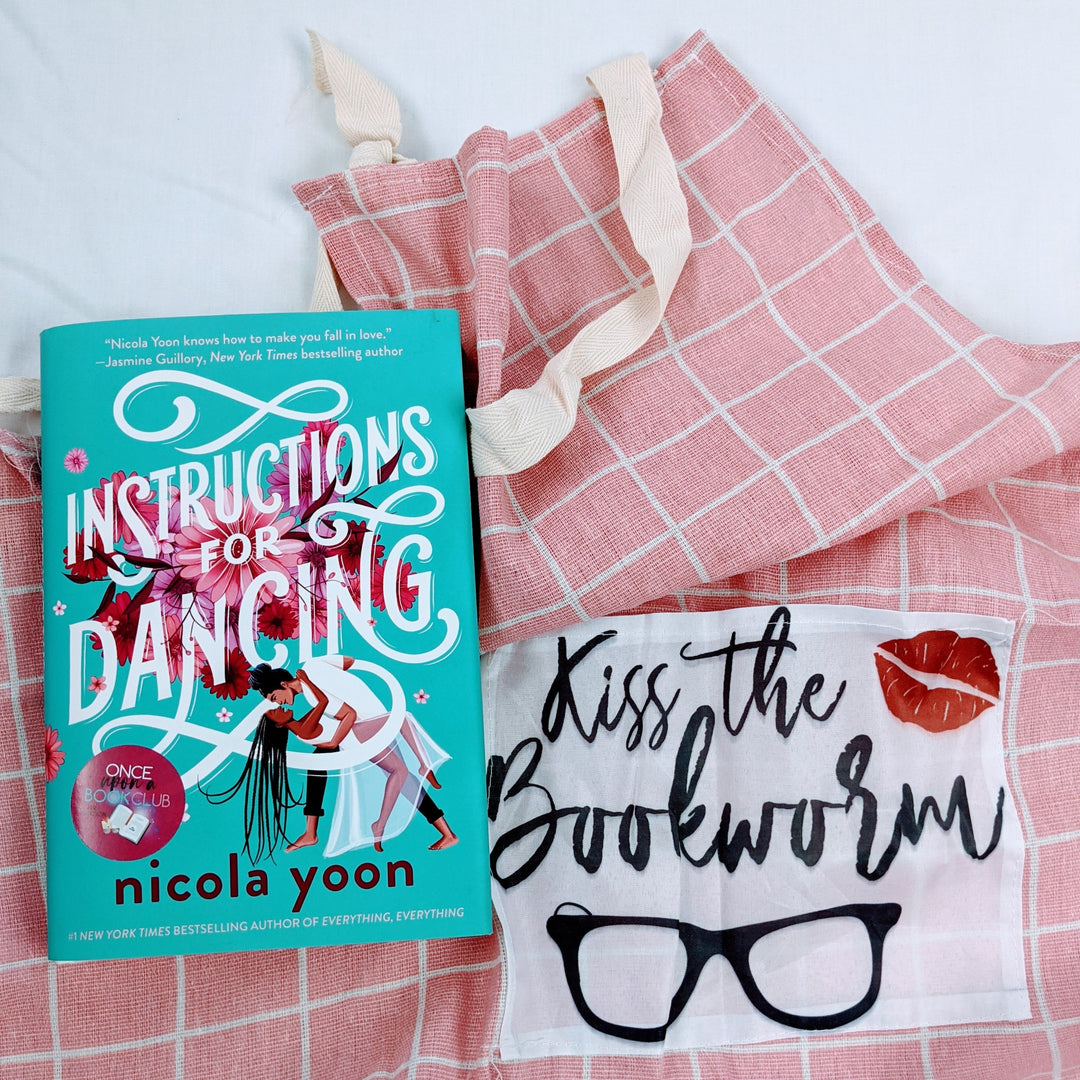 A copy of Instructions for Dancing by Nicola Yoon sits on a close up of the pink gingham apron featuring a pocket with the words "Kiss the Bookworm" and a pair of glasses and lipstick kiss mark. On a white background.