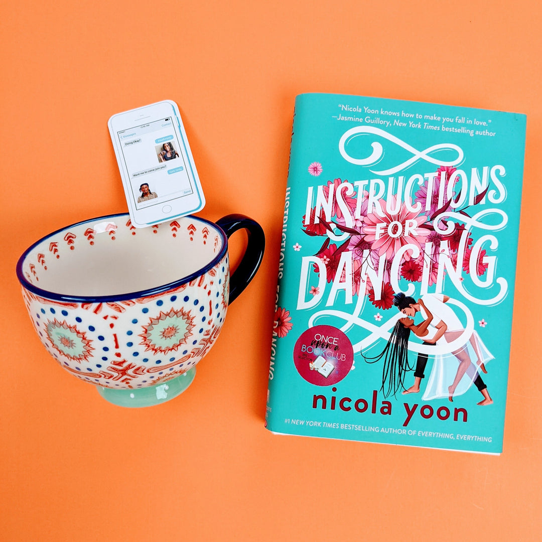An oversized mug painted with a dark blue, red, and light blue pattern sits next to a copy of Instructions for Dancing by Nicola Yoon. On an orange background.