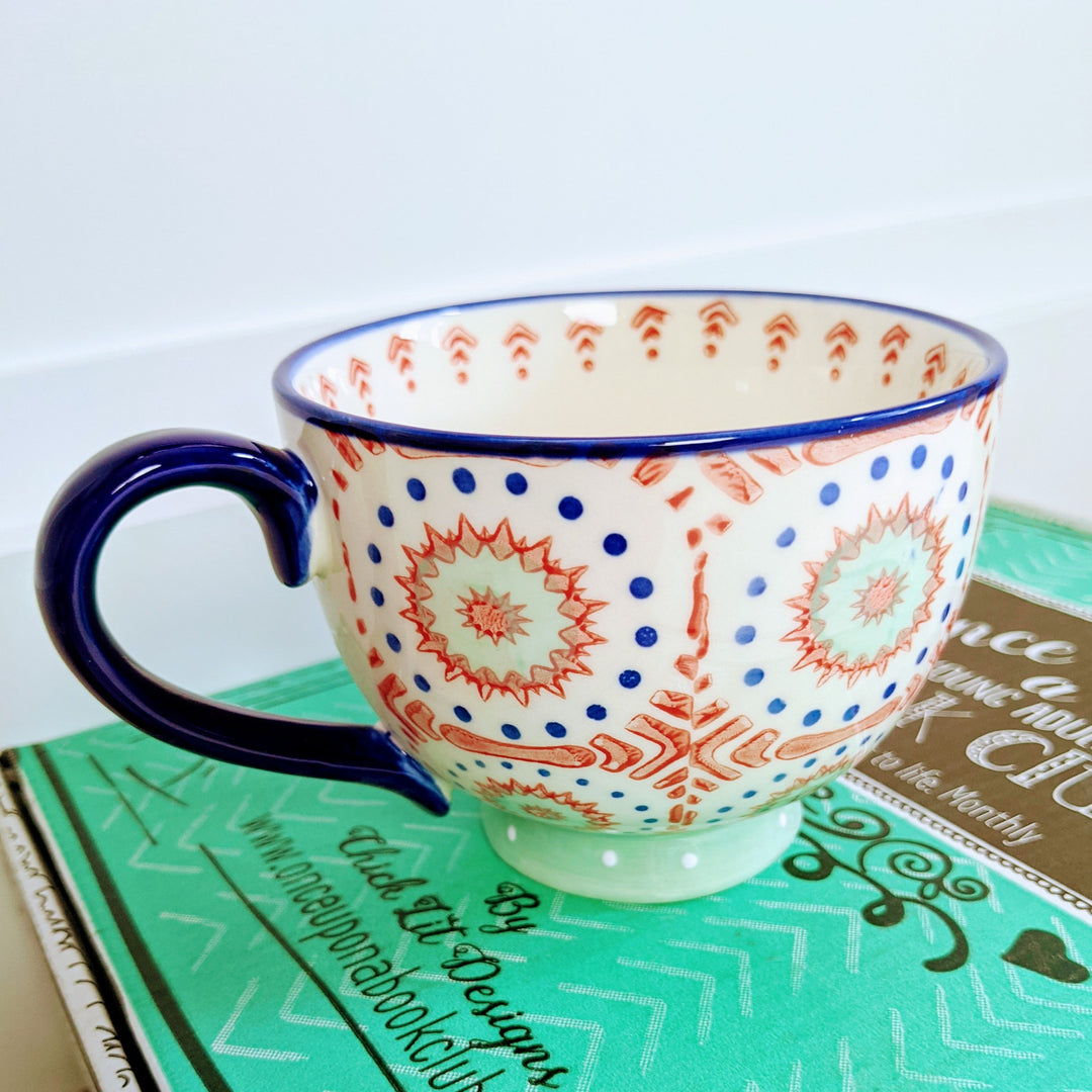 An oversized mug painted with a dark blue, red, and light blue pattern sits on a green OUABC box.