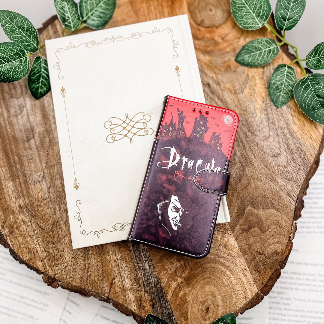 The front of a phone case inspired by Dracula by Bram Stoker. The phone case sits on a white book cover on a wood slice surrounded by greenery.